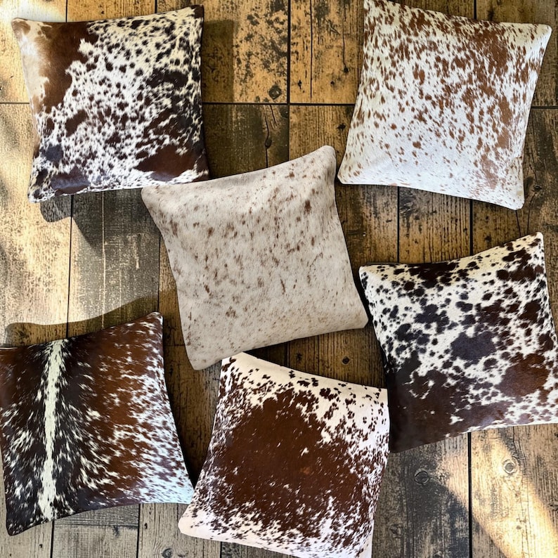 May include: Six square throw pillows featuring various cowhide patterns in shades of brown, white, and black. The collection includes spotted, speckled, and solid-toned animal print designs with a textured appearance arranged in a cluster.