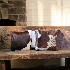 May include: Two square throw pillows featuring a cowhide pattern with patches of dark brown and white fur. The left pillow displays a large solid brown patch, while the right pillow shows a mottled brown and white spotted cowhide texture.