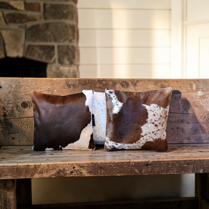 May include: Two square throw pillows featuring a cowhide pattern with patches of dark brown and white fur. The left pillow displays a large solid brown patch, while the right pillow shows a mottled brown and white spotted cowhide texture.