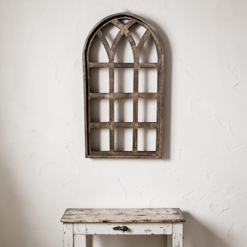 May include: A rustic, wooden wall decor piece in the shape of an arched window. The frame is made of weathered wood with a grid pattern, creating a vintage aesthetic. The piece is mounted on a white wall above a distressed, white table.