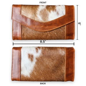 Op de afbeelding: Een bruin en witte koeienhuid portemonnee met een leren flap en afwerking. De portemonnee is 21,6 cm lang en 12,7 cm hoog. De voor- en achterkant van de portemonnee worden getoond. De portemonnee heeft een getextureerde uitstraling.
