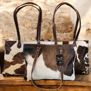 May include: Two brown and white cowhide tote bags with brown leather handles and straps. The larger bag has a zipper closure. The smaller bag has a tag that reads "Handmade in South Africa".