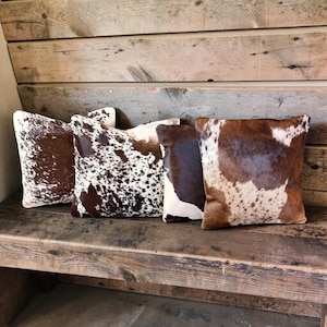 May include: Four square throw pillows featuring various cowhide patterns in shades of brown, white, and dark brown. The pillows display distinct natural animal hide textures with spotted, mottled, and solid color block designs arranged in a row.