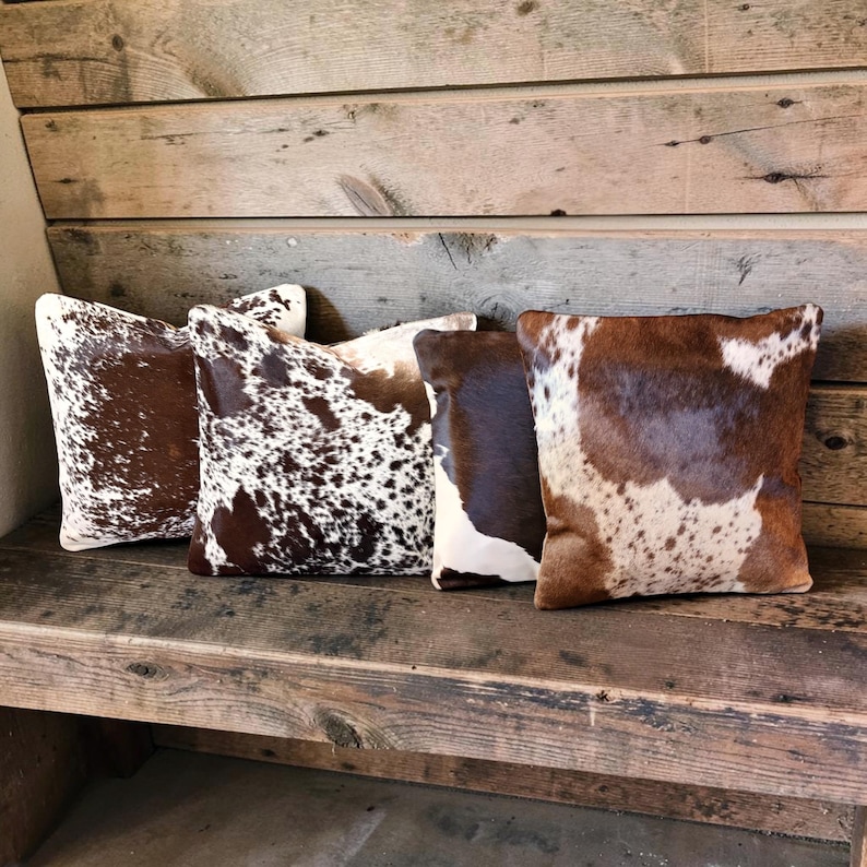 May include: Four square throw pillows featuring various cowhide patterns in shades of brown, white, and dark brown. The pillows display distinct natural animal hide textures with spotted, mottled, and solid color block designs arranged in a row.