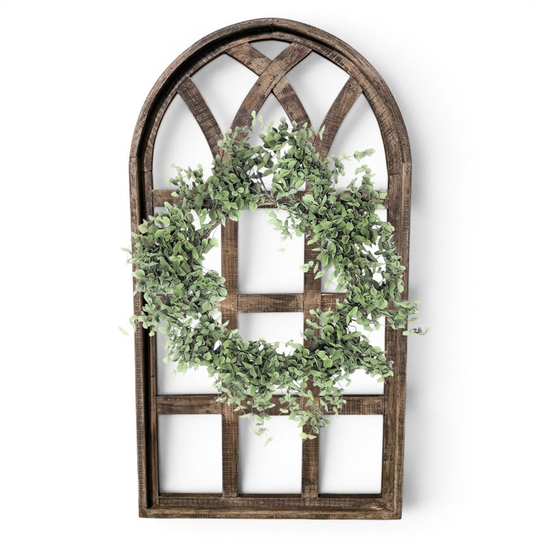 May include: A rustic, arched wooden window frame with a lattice design. A circular green leafy wreath is centered on the frame. The wood has a dark brown finish, and the wreath is a vibrant green, creating a natural, decorative piece.