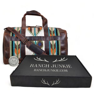 May include: A brown leather duffel bag with a colorful woven pattern. The bag has a shoulder strap and a zipper closure. The bag is sitting on a black box with the text "RANCH JUNKIE" and "RANCHJUNKIE.COM".