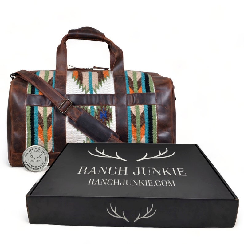 May include: A brown leather duffel bag with a colorful woven pattern. The bag has a shoulder strap and a zipper closure. The bag is sitting on a black box with the text "RANCH JUNKIE" and "RANCHJUNKIE.COM".