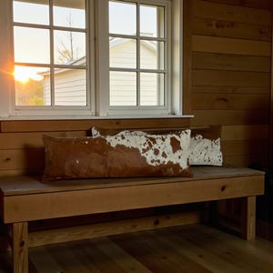 May include: Two rectangular cowhide pillows with brown and white patterns rest on a wooden bench. The bench is made of light-colored wood and is positioned near a window. The room has wood paneling and a view of a building outside.
