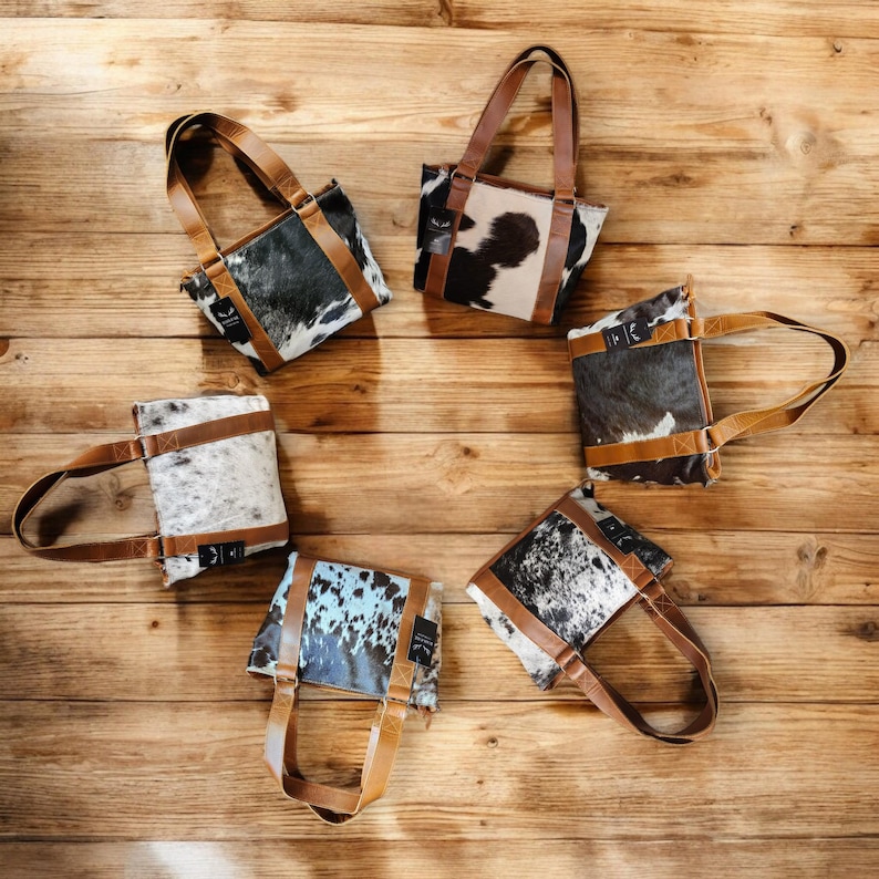 May include: Six small cowhide tote bags with brown leather handles arranged in a circle on a wooden surface. The bags are all different colors and patterns, including black and white, brown and white, and blue and white.