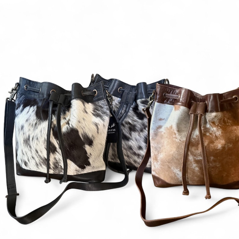 Puede incluir: Varios bolsos de cuero tipo cubo con estampados de piel de vaca. Los bolsos tienen bases de cuero negro, cordones y correas ajustables para el hombro. Los estampados de piel de vaca var&iacute;an en color, incluyendo blanco y negro, gris y marr&oacute;n.
