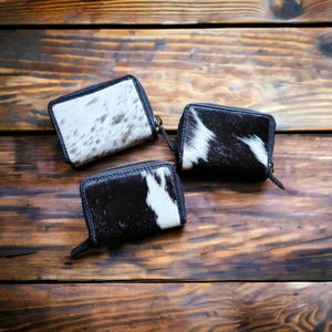 May include: Three small black leather wallets with a white and black cowhide pattern. The wallets are arranged on a wooden surface.