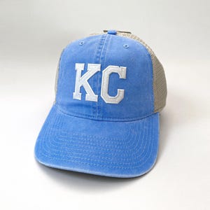 May include: A blue and tan trucker hat with white embroidered letters that spell "KC".