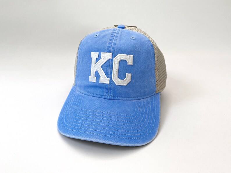 May include: A blue and tan trucker hat with white embroidered letters that spell "KC".