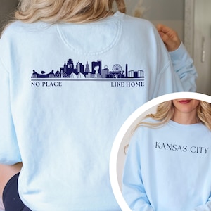 May include: Light blue sweatshirt featuring a navy blue skyline design with the text "NO PLACE LIKE HOME." The sweatshirt also has "KANSAS CITY" printed on the front. A comfortable, casual top.