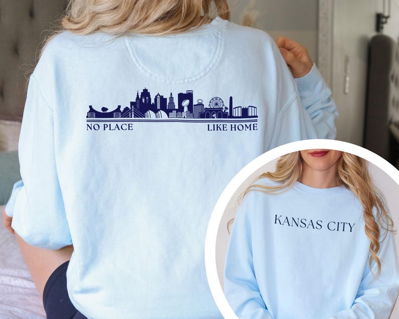 May include: Light blue sweatshirt featuring a navy blue skyline design with the text "NO PLACE LIKE HOME." The sweatshirt also has "KANSAS CITY" printed on the front. A comfortable, casual top.