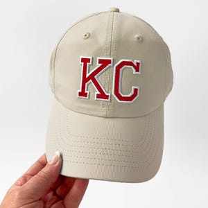 May include: A tan baseball cap with the letters "KC" embroidered in red with a white outline. The cap has a curved brim and a textured fabric. The cap is being held by a person.