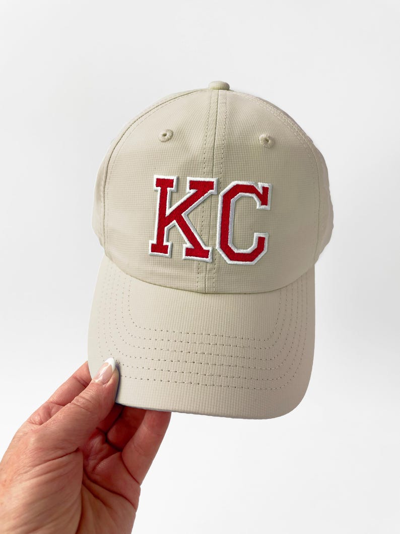 May include: A tan baseball cap with the letters "KC" embroidered in red with a white outline. The cap has a curved brim and a textured fabric. The cap is being held by a person.