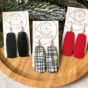 Op de afbeelding: Drie paar hangende oorbellen op witte kaartjes met de tekst "Kate Callahan Designs". De oorbellen zijn gemaakt van leer en hebben een zwart, rood en zwart-wit geruit ontwerp.