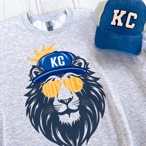 May include: A gray sweatshirt with a graphic of a lion wearing a blue baseball cap with the letters "KC" on it and sunglasses that say "Royals". The lion is wearing a gold crown. A blue mesh baseball cap with the letters "KC" on it is also in the image.