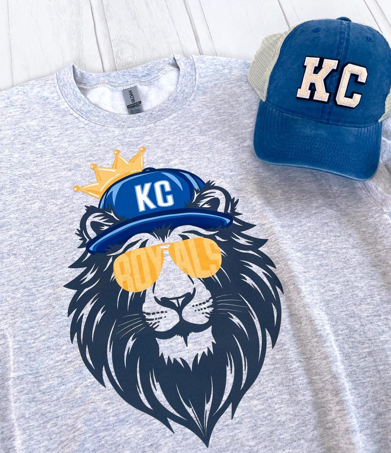 May include: A gray sweatshirt with a graphic of a lion wearing a blue baseball cap with the letters "KC" on it and sunglasses that say "Royals". The lion is wearing a gold crown. A blue mesh baseball cap with the letters "KC" on it is also in the image.