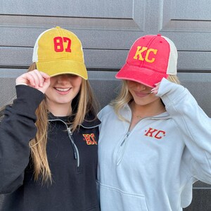 May include: Two people wearing trucker hats. One hat is yellow with the number 87 on it. The other hat is red with the letters KC on it. Both people are wearing sweatshirts with the letters KC on them.
