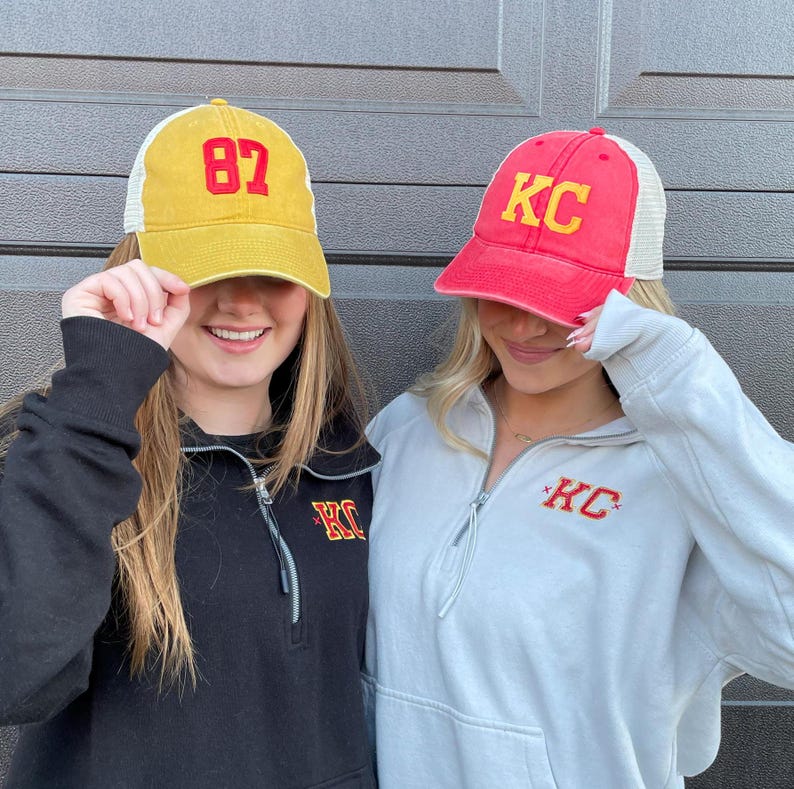 May include: Two people wearing trucker hats. One hat is yellow with the number 87 on it. The other hat is red with the letters KC on it. Both people are wearing sweatshirts with the letters KC on them.