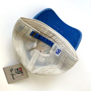 May include: A beige and blue mesh trucker hat with a blue denim brim and a white mesh back. The hat has a brown leather strap with a brass buckle and a white label with the text "The G Game" and a logo.