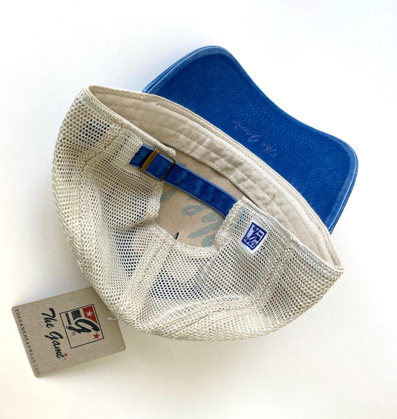 May include: A beige and blue mesh trucker hat with a blue denim brim and a white mesh back. The hat has a brown leather strap with a brass buckle and a white label with the text "The G Game" and a logo.