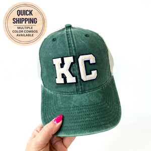 May include: A green and white trucker hat with the letters "KC" embroidered in white on the front.