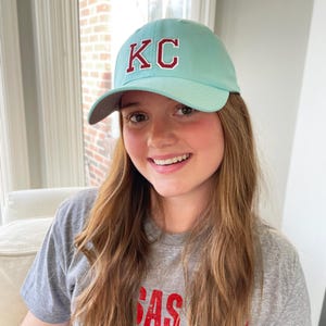 May include: A light blue baseball cap with the letters "KC" in red. The cap is worn with a gray t-shirt that has the word "SAS" in red. The image is taken indoors.