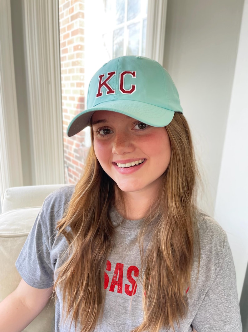 May include: A light blue baseball cap with the letters "KC" in red. The cap is worn with a gray t-shirt that has the word "SAS" in red. The image is taken indoors.