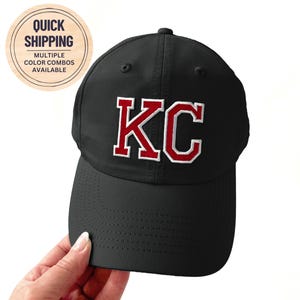 May include: Black baseball cap with the letters "KC" in red with a white outline. The cap has a curved brim and a structured crown. The image also includes a circular sticker with the text "Quick Shipping" and "Multiple Color Combos Available".