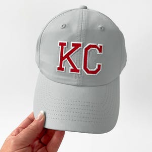 May include: A gray baseball cap with the letters "KC" in red with a white outline. The cap has a curved brim and a textured surface. The cap is a sports-themed accessory.