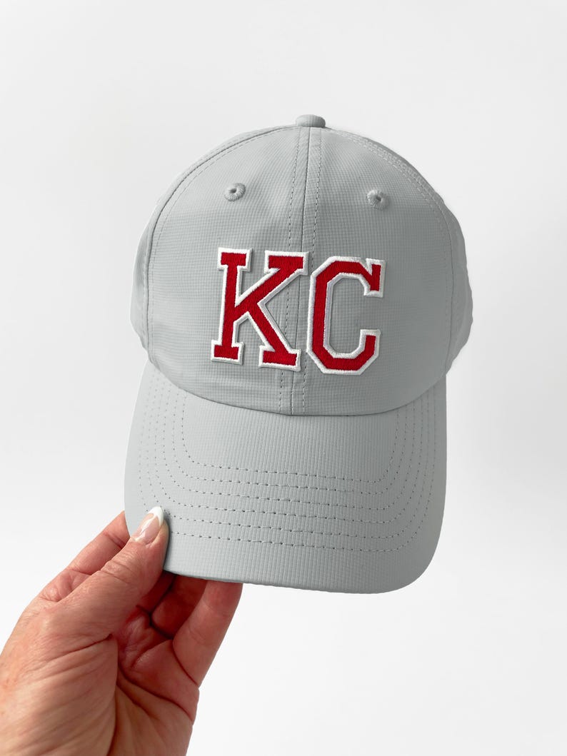 May include: A gray baseball cap with the letters "KC" in red with a white outline. The cap has a curved brim and a textured surface. The cap is a sports-themed accessory.