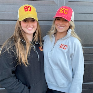 May include: Two women wearing baseball caps. The woman on the left is wearing a yellow cap with the number 87 on it. The woman on the right is wearing a red cap with the letters KC on it. Both women are wearing sweatshirts with the letters KC on them.