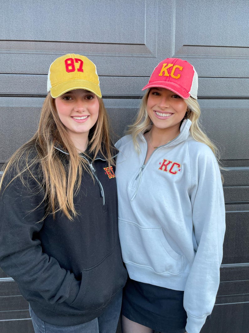 May include: Two women wearing baseball caps. The woman on the left is wearing a yellow cap with the number 87 on it. The woman on the right is wearing a red cap with the letters KC on it. Both women are wearing sweatshirts with the letters KC on them.