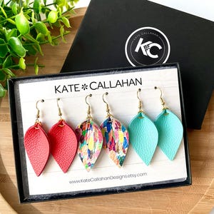 May include: Three pairs of leather earrings in a box. The earrings are in the shape of a teardrop and are available in red, turquoise, and a colorful abstract print.