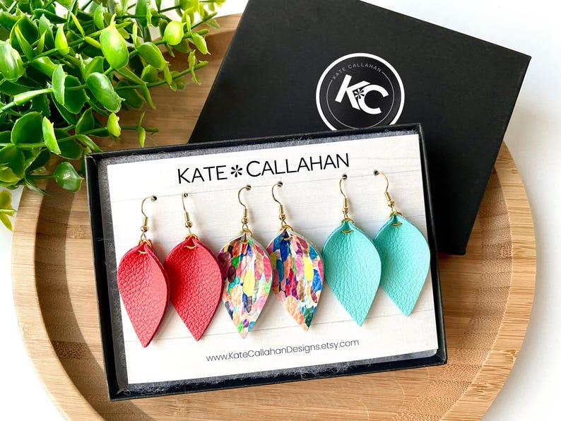 May include: Three pairs of leather earrings in a box. The earrings are in the shape of a teardrop and are available in red, turquoise, and a colorful abstract print.