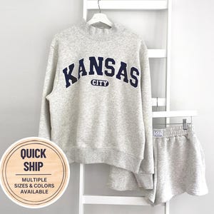 Puede incluir: Una sudadera de cuello alto en color gris jaspeado claro con el texto azul oscuro KANSAS CITY impreso en el pecho, combinada con pantalones cortos a juego en gris jaspeado claro con un pequeño logo KC en la cintura elástica, en una percha blanca.