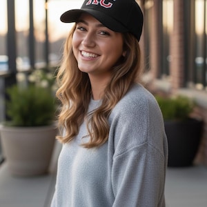 May include: Black baseball cap with the letters "KC" in red and white. The cap is worn by a person smiling at the camera. The person is wearing a light gray sweater.