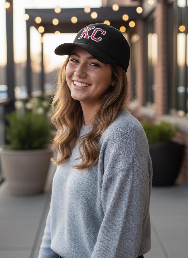 May include: Black baseball cap with the letters "KC" in red and white. The cap is worn by a person smiling at the camera. The person is wearing a light gray sweater.