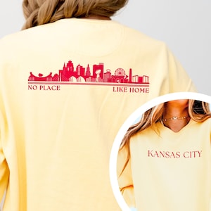 May include: A light yellow sweatshirt with a red skyline graphic and the text "NO PLACE LIKE HOME" on the back. The front of the sweatshirt has the text "KANSAS CITY" in red. The sweatshirt is a casual style.