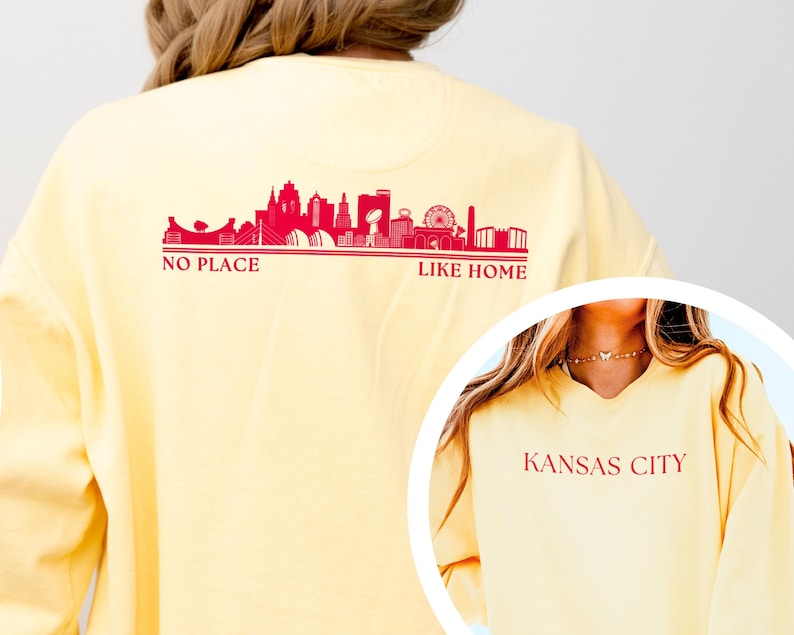 May include: A light yellow sweatshirt with a red skyline graphic and the text "NO PLACE LIKE HOME" on the back. The front of the sweatshirt has the text "KANSAS CITY" in red. The sweatshirt is a casual style.