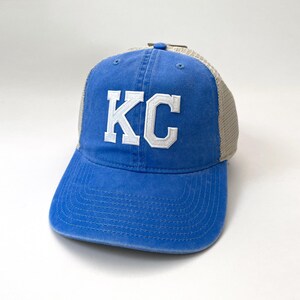 May include: A blue and beige trucker hat with a white embroidered "KC" on the front.