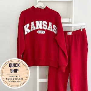 May include: A red matching set consisting of a mock neck sweatshirt and sweatpants. The sweatshirt features the white text KANSAS CITY in an arched font. The sweatpants have a small white patch with the letters KC on the elastic waistband.