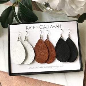May include: Three pairs of teardrop-shaped leather earrings in white, brown, and black. The earrings are displayed in a box with the text "KATE CALLAHAN" and a website address.