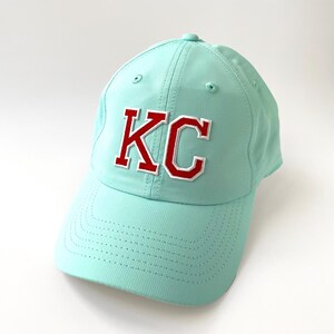 May include: A light teal baseball cap with the letters "KC" embroidered on the front in red with a white outline. The cap has a curved brim and a structured crown.