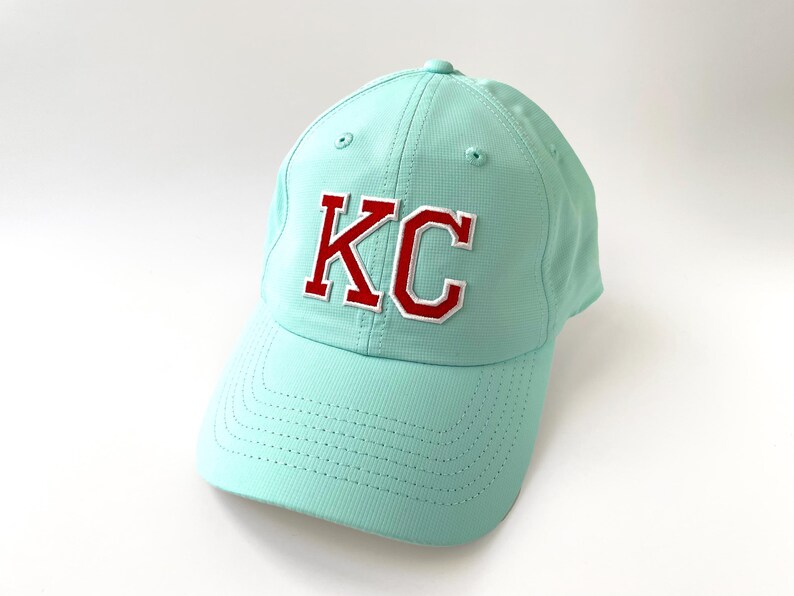 May include: A light teal baseball cap with the letters "KC" embroidered on the front in red with a white outline. The cap has a curved brim and a structured crown.