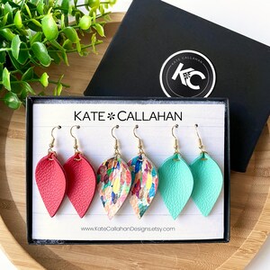 May include: Three pairs of leather earrings in a black box. The earrings are in the shapes of teardrops. One pair is red, one pair is turquoise, and one pair is a multi-colored abstract pattern. The box has the text "KATE*CALLAHAN" and "www.KateCallaghanDesigns.etsy.com" printed on it.