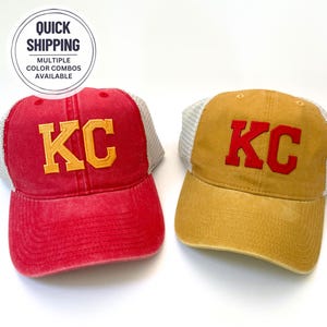 May include: Two trucker hats with a mesh back. One hat is red with a yellow "KC" embroidered on the front. The other hat is yellow with a red "KC" embroidered on the front.